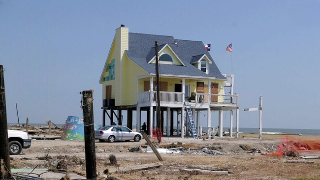 A two-story yellow house elevated 14 feet on thick wooden columns, standing entirely alone on a flat, scoured coastline.